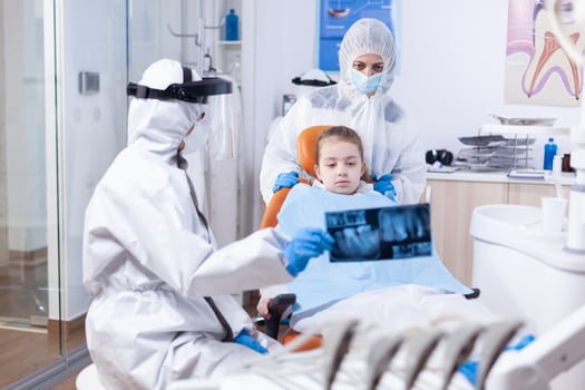 Dentist with face shield and ppe suit in dental clinic discussing about caries using radiography. Stomatolog in protectie suit for coroanvirus as safety precaution holding child teeth x-ray during consultation. photo