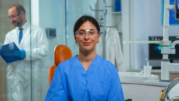 Portrait of smiling nurse in dental office while doctor is talking with patient in background. Stomatologist assistant looking on webcam sitting on chair in stomatological clinic. photo