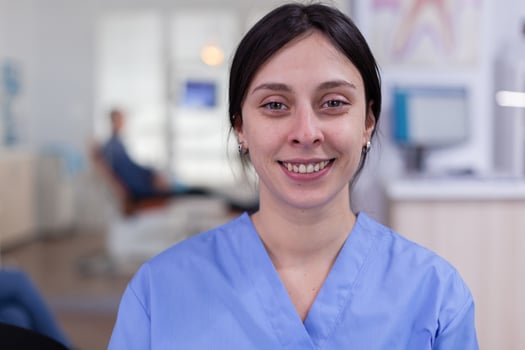Dental assistant looking at camera while doctor examining patient in background. Professional stomatologist nurse smiling on webcam sitting on chair in waiting room of stomatological clinic. photo