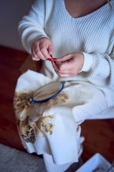 midsize woman in light clothes embroiders a picture in gold and beige tones while sitting on a white chair in a light room photo