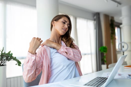 Portrait of young stressed woman sitting at home office desk in front of laptop, touching aching shoulder with pained expression, suffering from shoulder ache after working on laptop photo