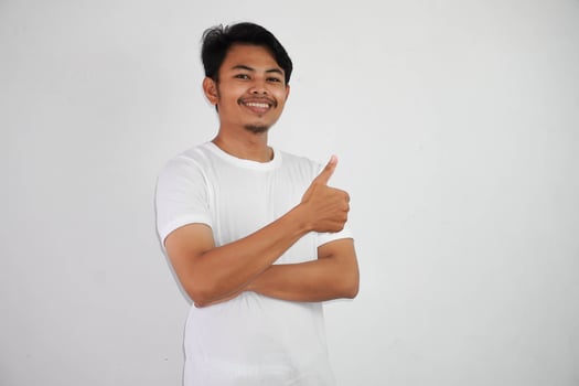 Portrait of cheerful asian man in wearing white t shirt smiling and showing thumbs up at camera isolated over white background photo