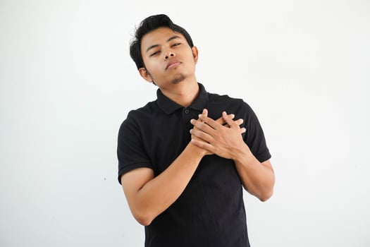 young asian man posing on a white backdrop laughing keeping hands on heart, concept of happiness. wearing black polo t shirt. photo