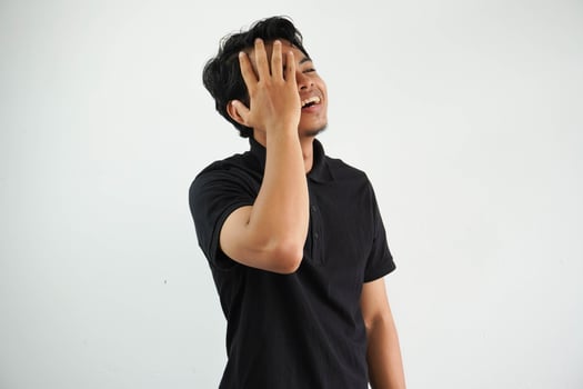 young asian man posing on a white backdrop having fun covering half of face with palm, wearing black polo t shirt. photo