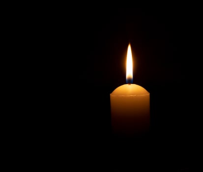 Single burning candle flame or light glowing on a big white candle on black or dark background on table in church for Christmas, funeral or memorial service with copy space. photo