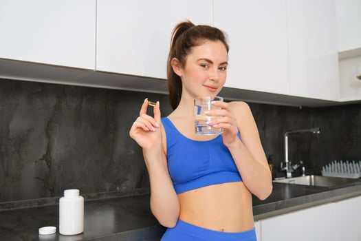 Portrait of healthy, smiling fitness woman, showing vitamins, fish oil dietary supplement for strong, fit body, drinking water with tablet, standing in ktichen photo