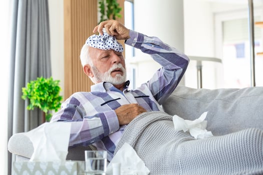 Mature man feeling sick with cold and fever at home, ill with flu disease sitting on the sofa with ice pack on his head photo