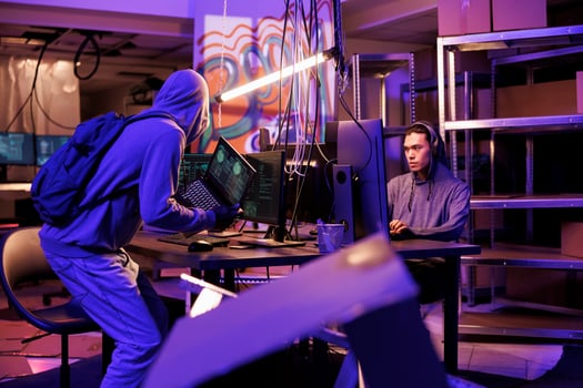 Hacker in hood bringing laptop with malware code running on screen to hideout place. Criminals planning crime and preparing cyberattack on server in abandoned warehouse at night photo
