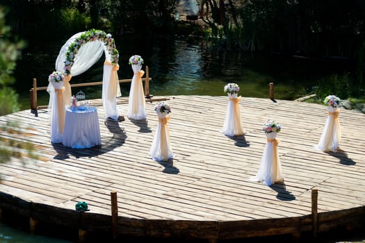 Wedding arch beautifully decorated and a small table, bouquets of flowers prepared for ceremony outdoors. Wooden surface in the form of a circle over the river for wedding event. photo