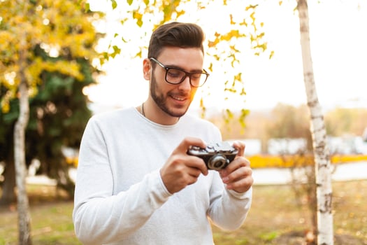 Young handsome hipster guy walks in a beautiful autumn park on the background of yellow leaves in warm sunny weather and takes pictures on a pig film camera. Autumn leisure time. Creative youth photo