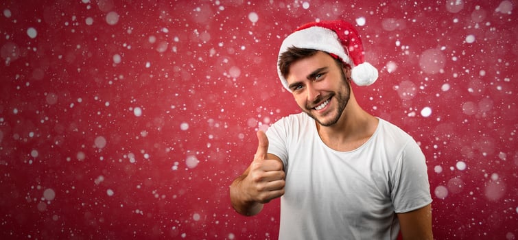 Young handsome caucasian guy in a white t-shirt and Santa hats stands on red background in studio and showing thumbs up. photo