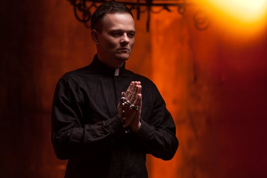 Young Catholic Praying priest. Portrait of priest Next to the candles prays with his hands folded near the face. Red wall on the background. Indoor handsome portrait of religion man photo