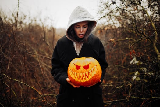 Happy Halloween Portrait of black modern lifestyle witch in hood and black coat mantle on wooden bush forest background. Holding pumpkin in hands photo