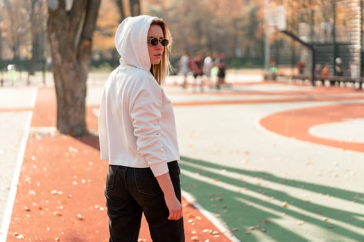 Outdoor close up portrait of young beautiful woman with in glasses, dressed in a white sweater, near the sportsground. Woman in a hood turned her head to the camera photo
