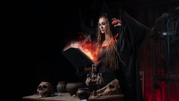 Halloween concept. Witch dressed black hood with dreadlocks standing dark dungeon room use magic book for conjuring magic spell photo