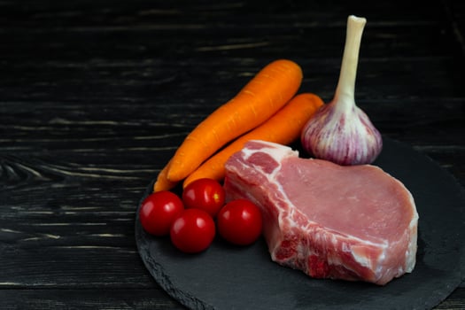 Top view of one pieces raw pork chop steaks with cherry tomatoes carrot and garlic on a black stone cutting board. photo