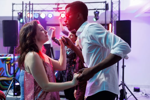 Romantic young couple holding hands and dancing together while clubbing. Diverse boyfriend and girlfriend partying and moving on dancefloor in nightclub illuminated with lights photo