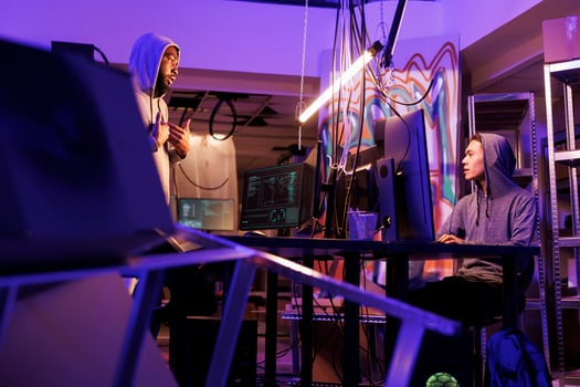 Two diverse hackers working together to hack computer system in abandoned warehouse. Asian and african american internet criminals discussing malicious software code in dark workspace photo