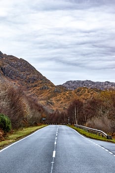 Empty rural road leading through a scenic autumn landscape with colorful foliage and mountain backdrop under a cloudy sky. photo
