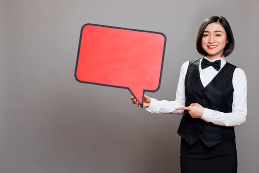 Smiling asian waitress in uniform holding red empty chat bubble and pointing with finger portrait. Cheerful woman receptionist showing blank message banner and looking at camera photo