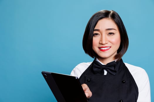 Smiling cafe waitress holding digital tablet at work and looking at camera with cheerful facial expression. Joyful cafe woman worker using portable gadget portrait on blue background photo
