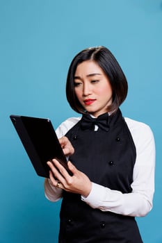 Restaurant young asian woman employee wearing uniform tapping on digital tablet. Cafeteria attractive waitress answering customer online message on portable gadget in studio photo