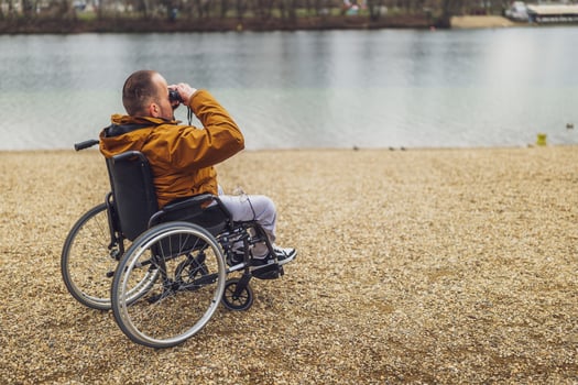 Paraplegic handicapped man in wheelchair is using binoculars outdoor. He is watching nature by the lake. photo