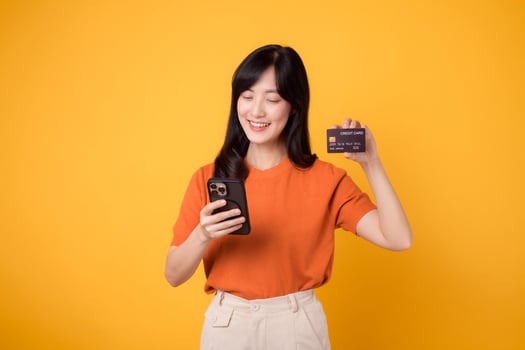 Enthusiastic Asian woman in her 30s, using smartphone and showing credit card on yellow background. Swift online shopping. photo