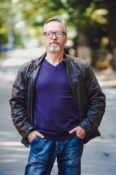 Handsome middle age man with gray hair in an outdoor setting. Leather jacket. Blue sweater. Eyeglasses. Bearded confident businessman. Blurred background. Hands in pockets. photo