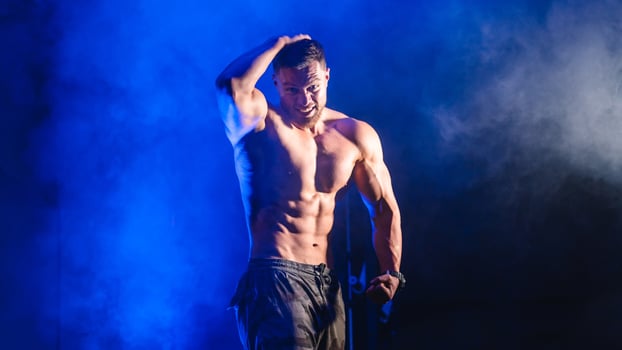 Bodybuilder showing belly and biceps muscles. Strong man flexing muscles. Fitness model posing at camera. Black and blue light background. Athlete looking straight at camera photo