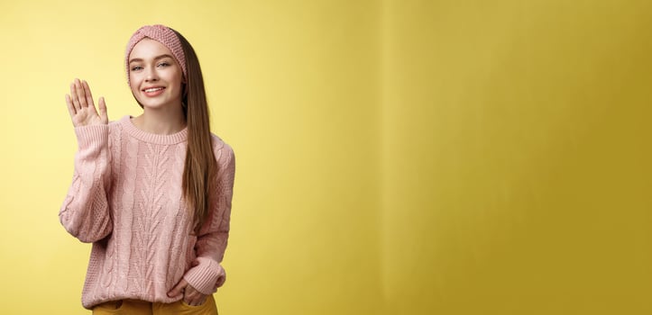 Hello adventures. Charming glamour 20s girl in sweater, knitted headband waving palm saying hi friendly smiling, holding hand in pocket gretting friends, posing positive and carefree over yellow wall photo