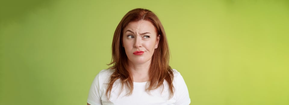 Perplexed doubtful unsure redhead wife smirking unsatisfied frowning look aside displeased disappointed hesitating pondering strange advice thoughtfully stare away stand green background photo