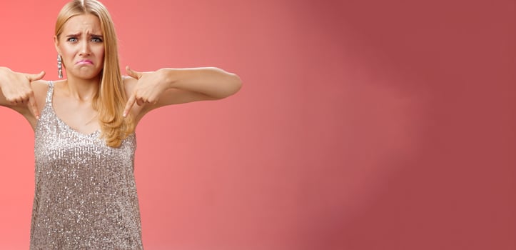 Whining complaining upset depressed blond woman in silver dress crying boyfriend broke-up prom night standing unhappy expressing sorrow regret disappointment pointing down red background photo