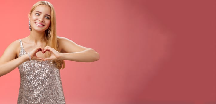 Romantic passionate charming lovely blond glamour woman in silver dress brilliants show heart love gesture express sympathy passion, adore relationship smiling delighted, standing red background photo