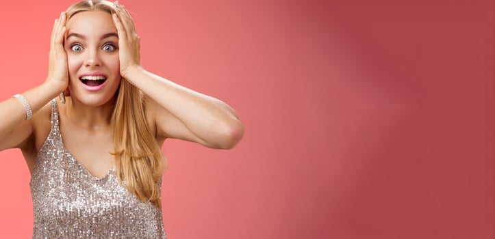 Amused surprised excited young blond attractive woman in silver glittering stylish dress hold head hands widen eyes speechless look camera unbelievable thrill fan see famous person, red background photo