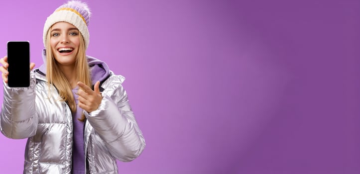 Satisfied amused good-looking blond girl suggest take look smartphone display smiling happily pointing mobile phone delighted talking about awesome new app features, standing purple background photo