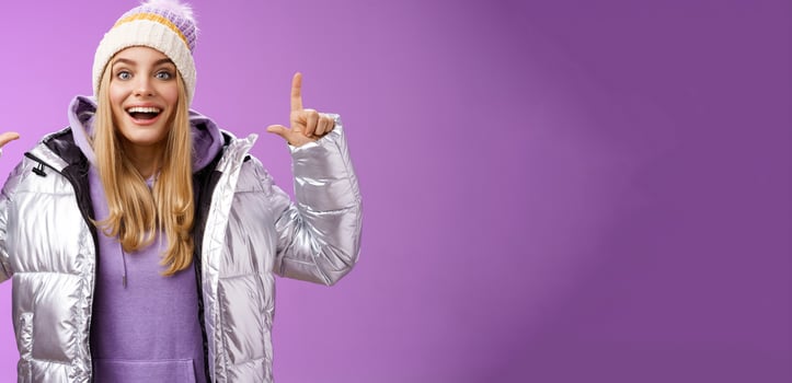 Excited carefree cheerful fair-haired european girl in silver jacket winter hat raising hands pointing up have excellent idea smiling broadly speaking passionately standing purple background photo