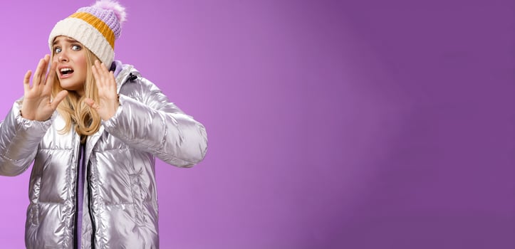 Afraid intense uncomfortable young fair-haired woman in winter hat silver stylish dress run away raise hands self-defense turn head away scared friend spill drink new outfit, purple background photo