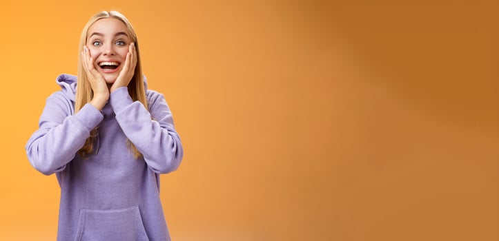 Surprised happy astonished fair-haired girlfriend receive incredible super cute gift touch cheeks amused reacting satisfied impressed standing excited orange background in casual hoodie photo