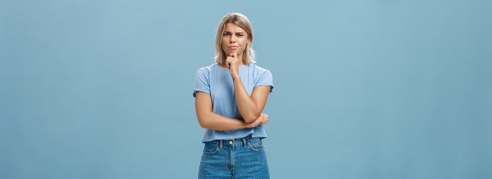 I doubt it good idea. Suspicious intense creative young female coworker in outdoor outfit frowning from doubtful thoughts holding hand on chin while thinking expressing disbelief over blue wall photo