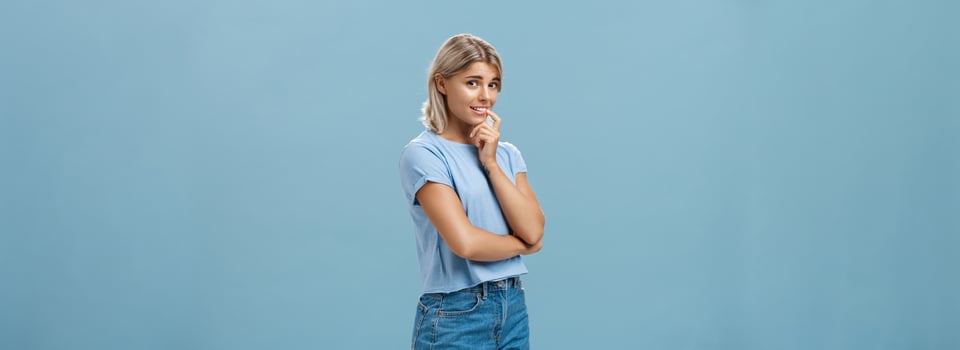 Studio shot of unconfident silly and insecure cute feminine girl with blond hair standing half-turned over blue background holding finger on lip while being shy ask question smiling hesitating photo