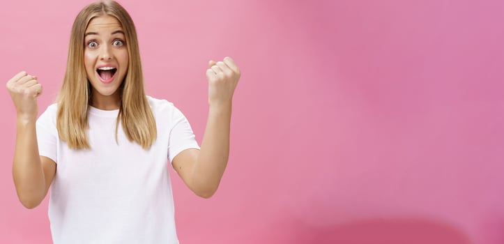 Excited cheerful and optimistic charming woman with fair hair in white t-shirt raising fists in victory and triumph yelling yes in success and amazement standing supportive against pink background photo