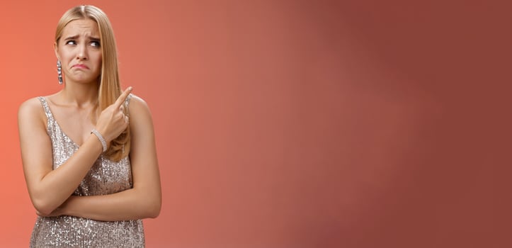 Upset doubtful unsure cute blond funny glamour girl in silver evening dress cringing grimacing hesitant pointing looking upper right corner suspicious, uncertain standing red background photo