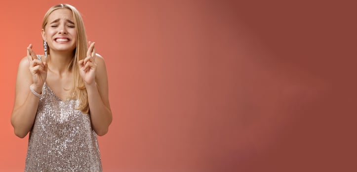 Nervous hopeful blond young woman in silver trendy dress close eyes grimacing eager wish come true cross fingers good luck praying supplicating anticiapting fortune, standing worried red background photo