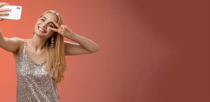Stylish fabulous glamour young blond woman in glittering silver dress tilting head carefree show peace gesture victory sign extend arm holding smartphone taking selfie recording video post online photo