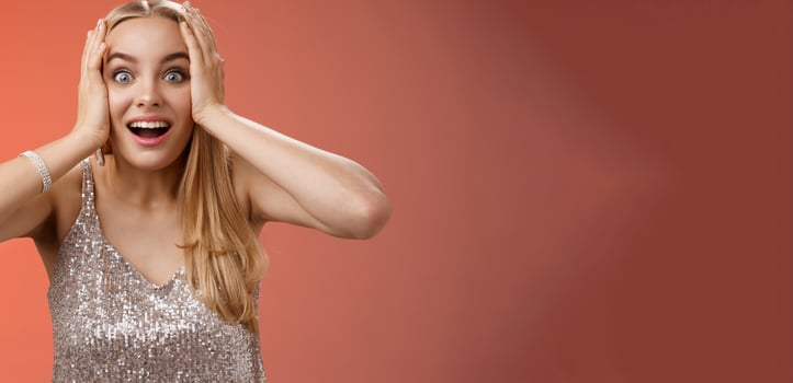 Amused surprised excited young blond attractive woman in silver glittering stylish dress hold head hands widen eyes speechless look camera unbelievable thrill fan see famous person, red background photo