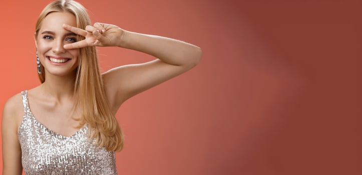 Joyful carefree attractive confident blond woman in stylish silver dress show disco sign victory peace gesture near eye smiling having fun enjoying awesome party grinning dancing red background photo