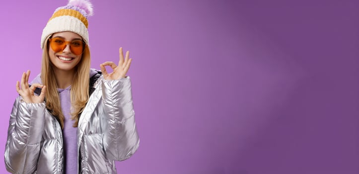 Everything fine thanks. Charming flirty blond confident woman in silver stylish jacket sunglasses winter hat show okay no problem ok gesture smiling affirmative, liking awesome day purple background photo