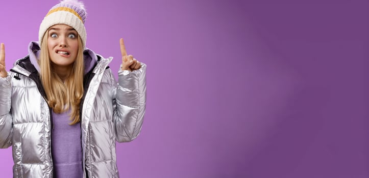 Scared unsure worried awkward young cute blond girl biting lip stooping pointing up widen eyes nervously scared someone find out secret standing embarrassed and intense purple background photo