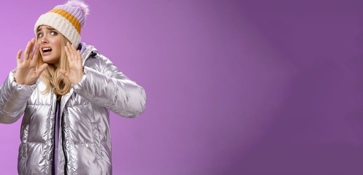 Afraid intense uncomfortable young fair-haired woman in winter hat silver stylish dress run away raise hands self-defense turn head away scared friend spill drink new outfit, purple background photo